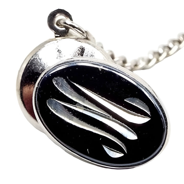 Front view of the Mid Century vintage faux hematite tie tack. The metal is silver tone in color. The tie tack is oval and has a dark metallic gray imitation hemtatite front with a curvy script initial M in the middle. The back clutch has a chain with a bar on the end.