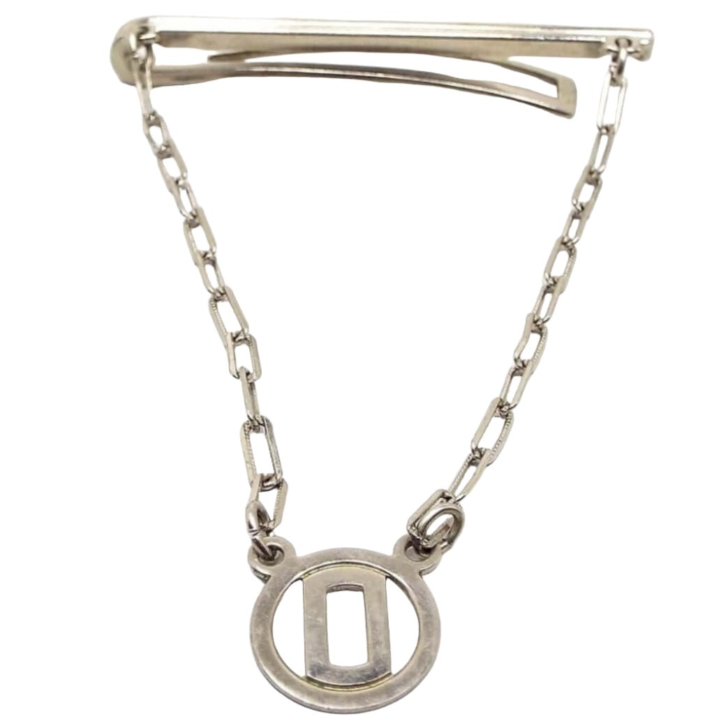 Front view of the Mid Century vintage tie bar chain. The metal is silver tone in color. The top bar has a very thin line design on the front and a curved back bar with open middle. The back bar is cuved outwards some, so it would be better for thicker style ties. Small cable chain with rectangle links comes down from each side of the bar and there is a round open charm at the bottom with a block letter initial O in the middle.