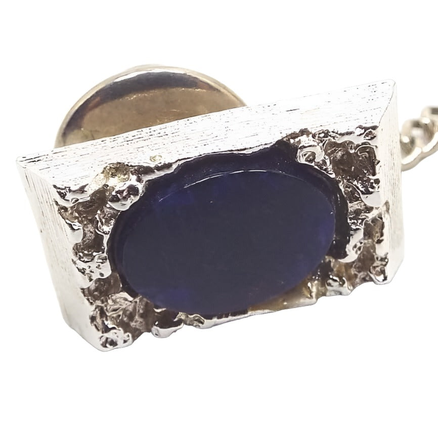 Front view of the Mid Century vintage sodalite tie tack. The dark blue sodalite gemstone cab is oval in shape and is surrounded by a nugget style textured silver tone rectangle setting. The clutch on the back has a chain with a small bar on the end.