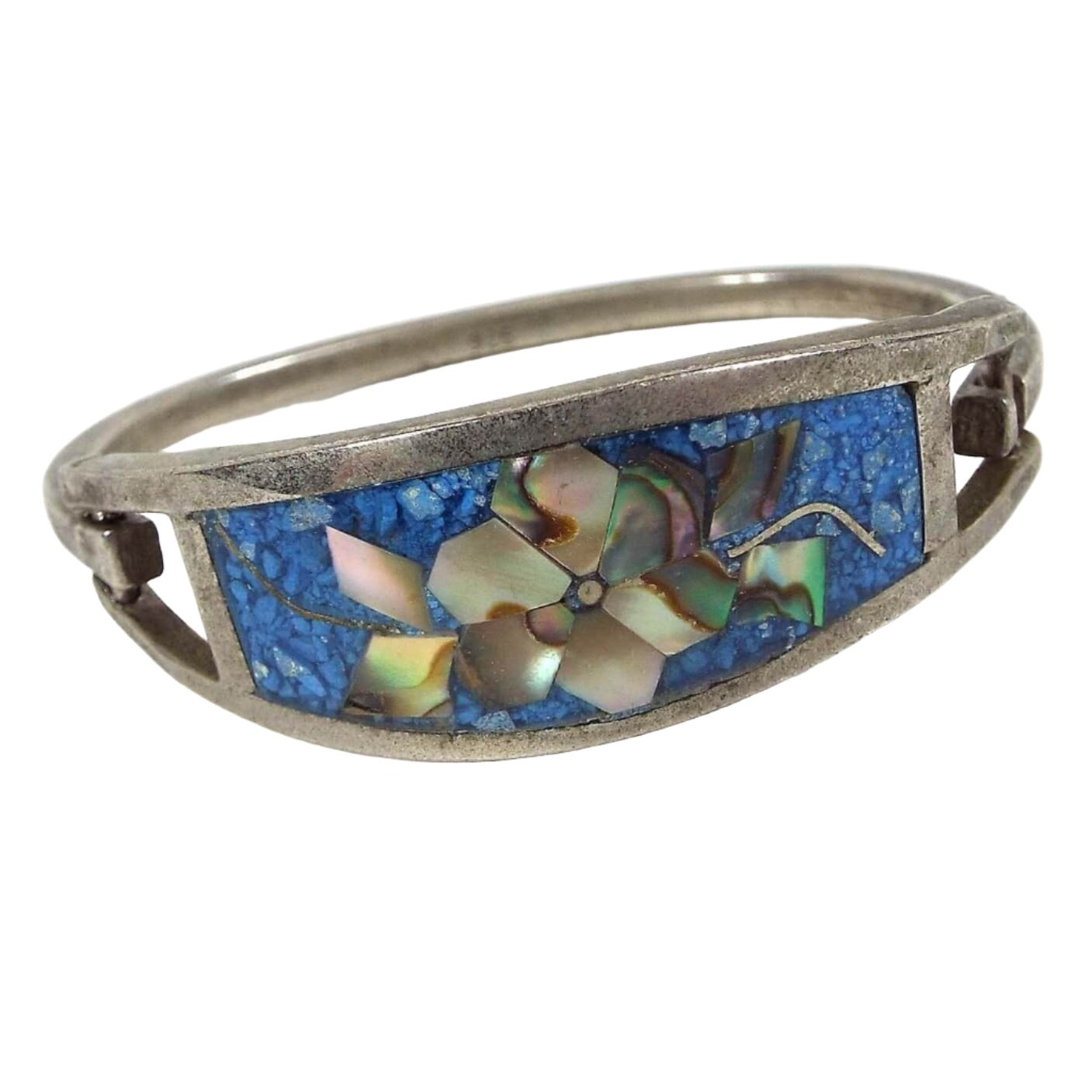 Angled front and side view of the retro vintage Mexican hinged bangle bracelet. The front has a large curved area with resin and tiny blue dyed stone chips. The middle has a flower design with pieces of multi color pearly inlaid abalone shell making up the petals and leaves on either side. The metal is silver tone in color and has a curved band that is hinged to one side of the top piece and has a slight hook on the other to open and close the bracelet.