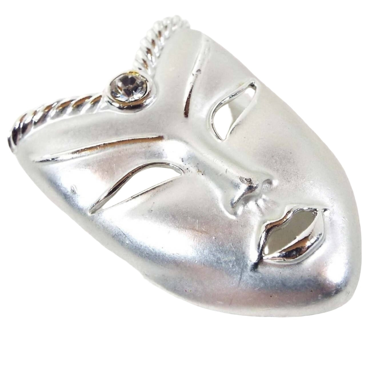 Front view of the retro vintage mask brooch pin. The metal is mostly matte silver tone in color with shiny silver tone color accents around the open cut out facial features. The top part angle's inward like a V shape and has a single round clear rhinestone in the middle.