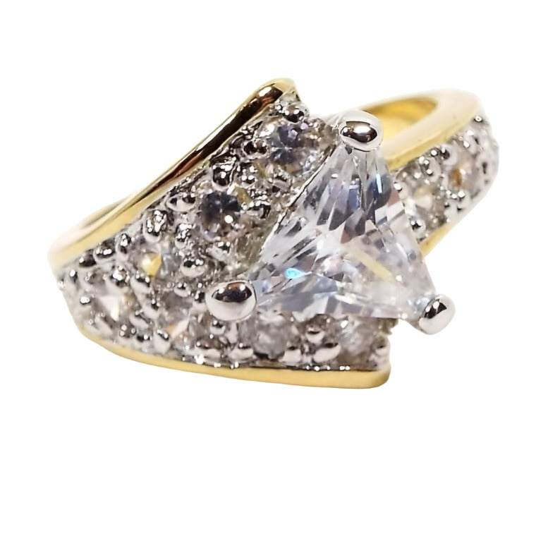 Front view of the retro vintage Edco cubic zirconia cocktail ring. The band and setting is gold tone in color. Ring has a nice asymmetrical design. One side of the top has a wide flared triangle style shape with small round pavé style prong set CZ stones. The other side has a single row of prong set CZ stones. On top is a larger triangle shaped trillion cut CZ stone.