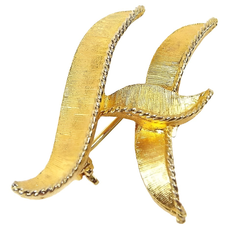 Front view of the Mid Century vintage letter brooch pin. The metal is matte brushed textured gold tone in color. It has a wavy block initial design of the letter H.