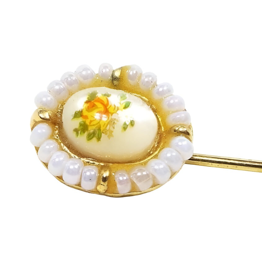 Front view of the top of the Mid Century vintage floral stickpin. The metal is gold tone in color. The top part is oval in shape and has a domed mother of pearl shell cab in the middle. There is a decal on top of the mother of pearl with a flower design in yellow and orange with green leaves. The outer edge of the oval has small pearly glass seed beads with small gold tone beads at each side.