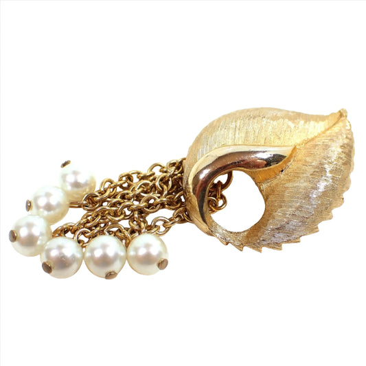 Front view of the Mid Century vintage tassel brooch pin. The brooch is shaped like an open leaf and is gold tone plated in color. The metal is matte brushed textured except for an area by a cut out in the middle that has shiny gold tone metal. There is a chain tassel at the end with white plastic faux pearls at the end of each chain.