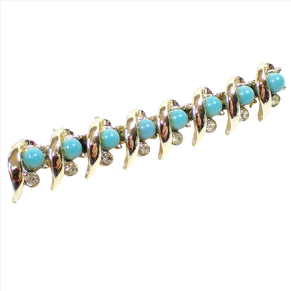 Angled enlarged front view of the Mid Century vintage bar brooch pin. The metal is gold tone plated in color. There are long leaf like shapes of metal that curve across the front. In between those are domed round plastic cabs in a turquoise blue color. Underneath each blue cab is a small round clear rhinestone.