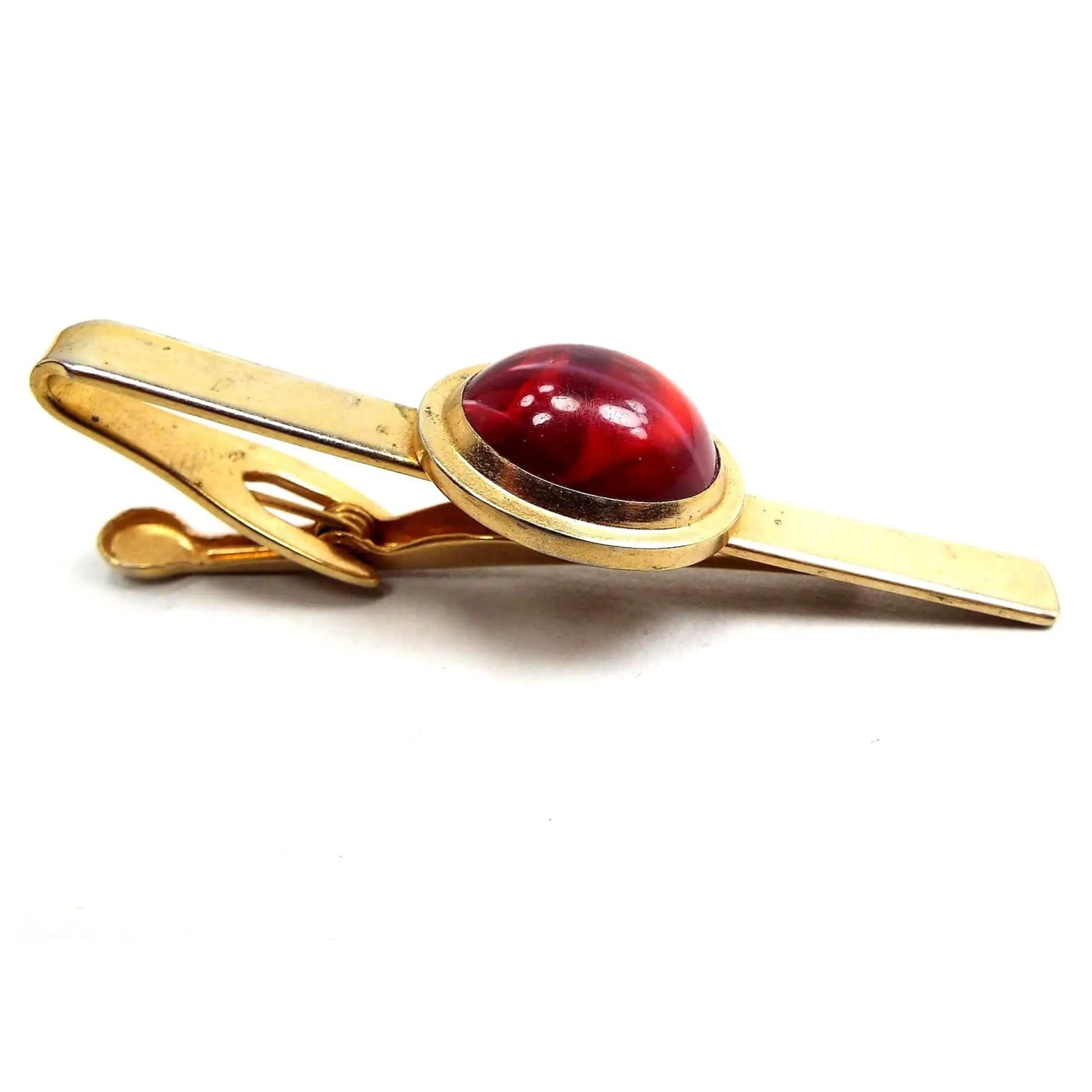 Angled view of the Mid Century vintage Pioneer lucite tie clip. The metal is gold tone in color. There is an oval in the middle with a marbled red domed lucite cab.