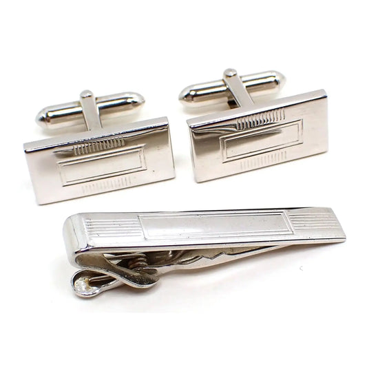 Enlarged view of the Mid Century vintage Swank mens jewelry set. The metal is silver tone in color. There is a pair of rectangle cufflinks at the top with rectangle and lines design in the middle. Below the cufflinks is a matching tie clip.