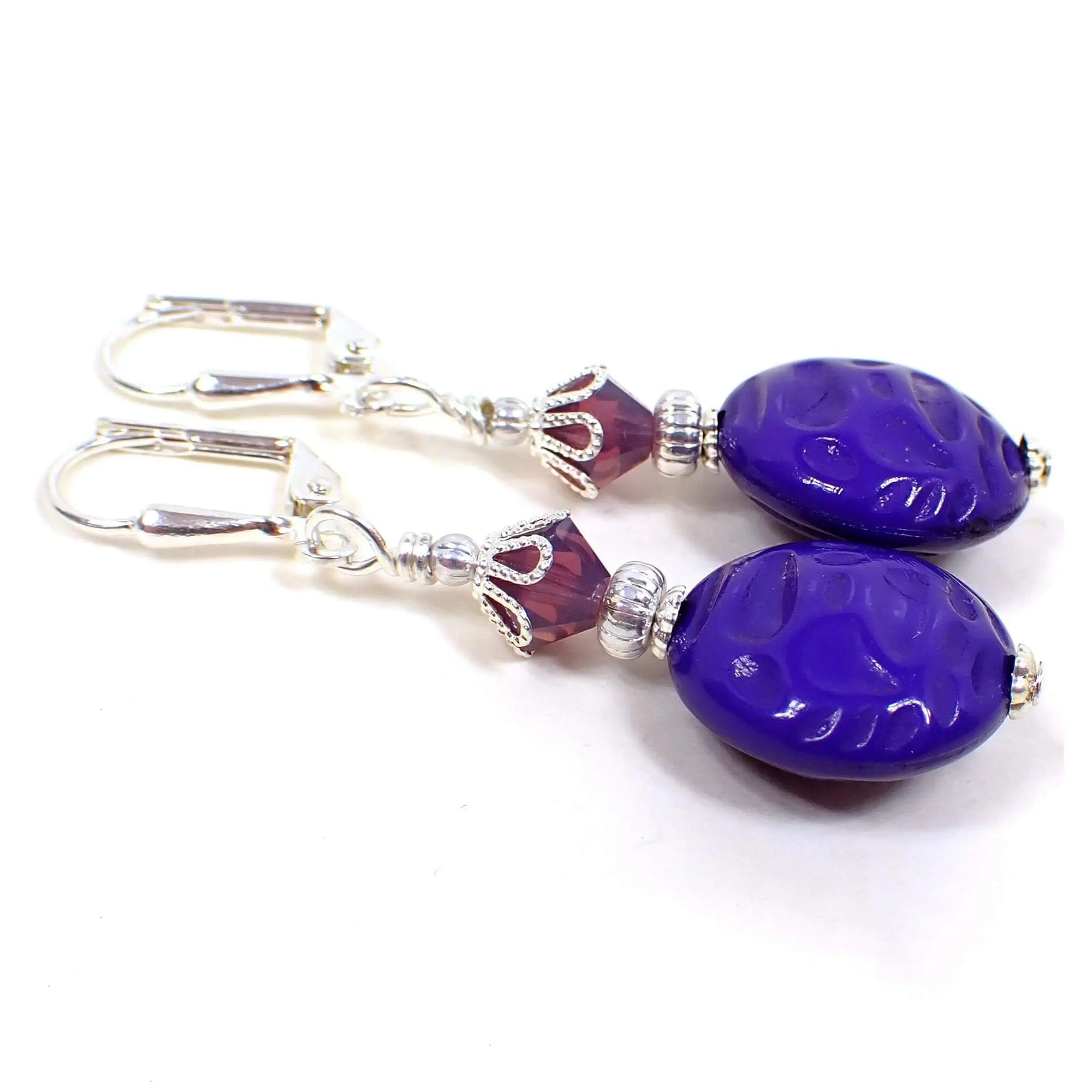 Side view of the handmade drop earrings. The metal is silver plated in color. There are purple semi translucent faceted glass crystal beads at the top. The bottom beads are vintage lucite beads that are puffy round shaped with an indented pattern and are a bright bluish purple in color.