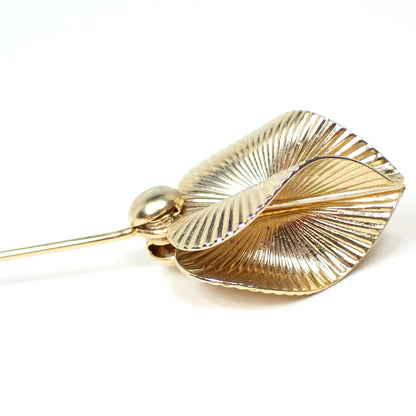 Enlarged top view of the retro vintage stick pin. The metal is gold tone in color. There are two curved and line textured leaf like designs at the top. 