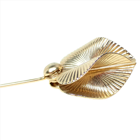 Enlarged top view of the retro vintage stick pin. The metal is gold tone in color. There are two curved and line textured leaf like designs at the top. 