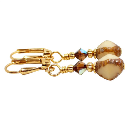 Enlarged side view of the small handmade drop earrings. The metal is gold plated in color. There are brown faceted glass beads at the top. The bottom Czech glass beads are bicone shaped and have brown with off white cut out areas on them.