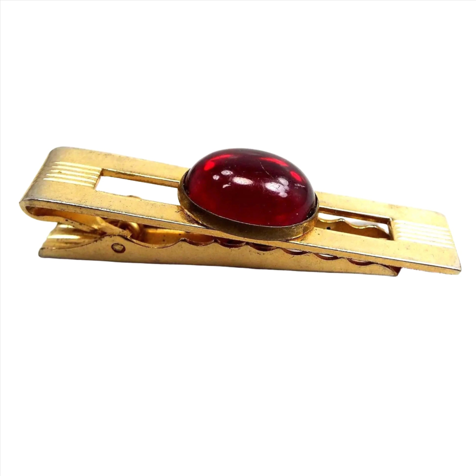 Angled side and front view of the Anson Mid Century vintage lucite tie clip. The metal is gold tone in color. There is an open cut out rectangle in the middle. Over that is an oval bezel set red lucite cab. There are etched lines on the metal on each end. The back has an alligator style clip.