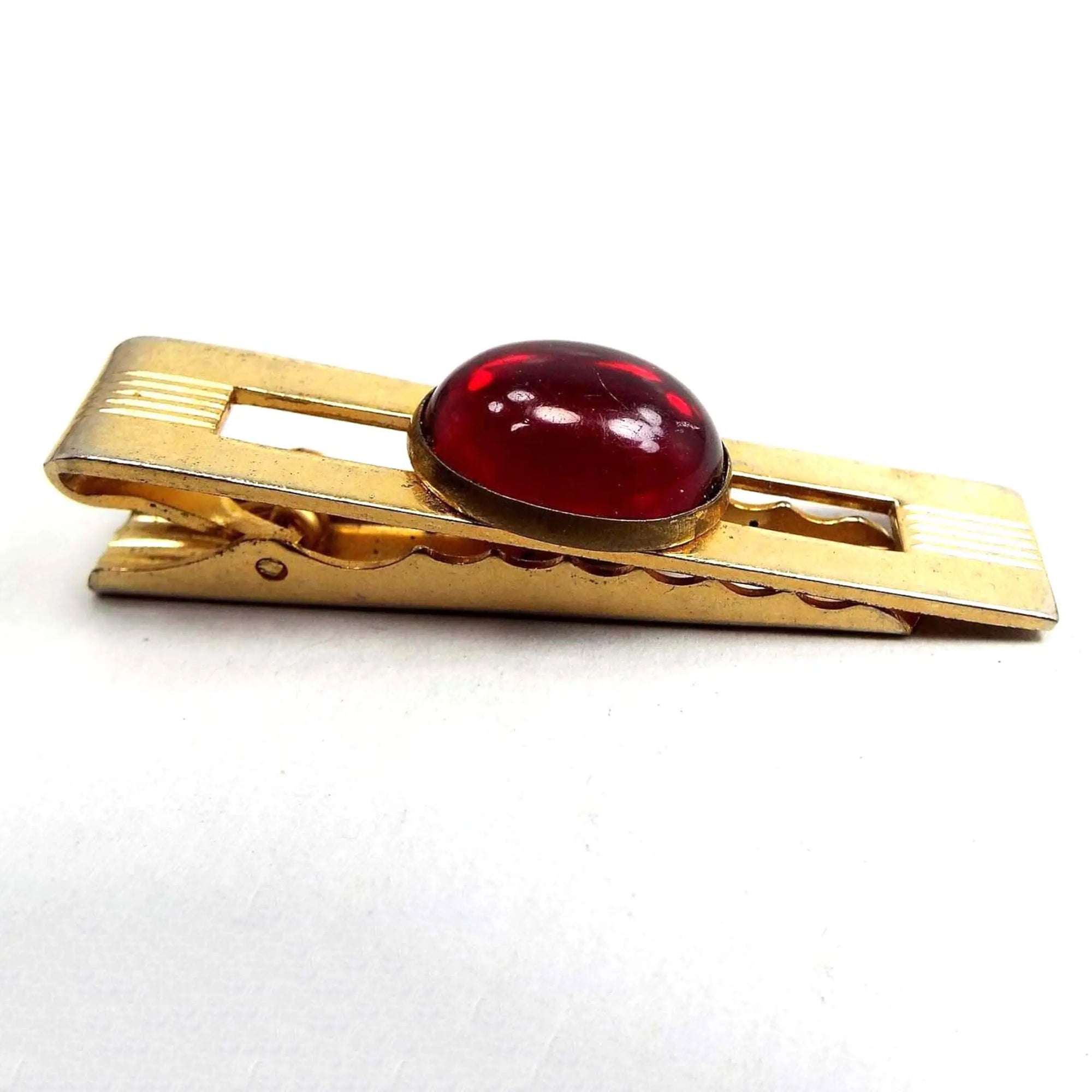 Angled side and front view of the Anson Mid Century vintage lucite tie clip. The metal is gold tone in color. There is an open cut out rectangle in the middle. Over that is an oval bezel set red lucite cab. There are etched lines on the metal on each end. The back has an alligator style clip.