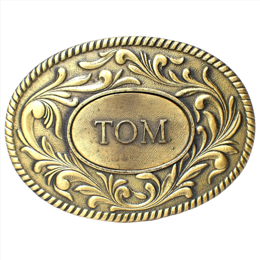 Front view of The Kinney Co retro vintage belt buckle from 1977. It is oval and antiqued brass in color. There is a raised leaf design around the edge. The middle has another oval with the name Tom in all capital block letters.