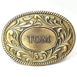 Front view of The Kinney Co retro vintage belt buckle from 1977. It is oval and antiqued brass in color. There is a raised leaf design around the edge. The middle has another oval with the name Tom in all capital block letters.