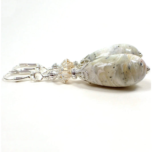 Side view of the large handmade earrings with vintage lucite beads. The metal is silver plated in color. There are light champagne color glass crystal beads at the top. The bottom lucite beads are plump teardrop shaped and have marbled light shades of gray, peach, pink, white, yellow, and small speckles here and there.