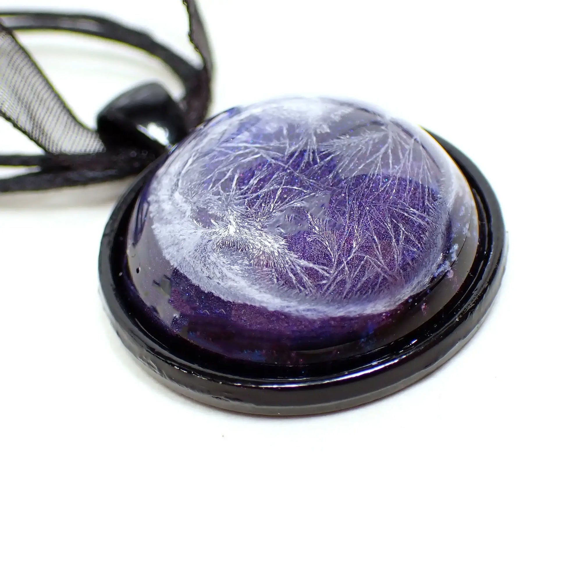 Enlarged view of the handmade resin pendant. The necklace part shows three black strands of faux leather cord and a strand of organza ribbon. There is a black coated round pendant with a domed resin cab. The resin has an iridescent purple background and an abstract crystalline like frost design in white through it.