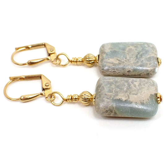 Top view of the handmade aqua terra jasper earrings. The metal is gold plated in color. There are two metal beads at the top of the earrings. The bottom stone bead is a manmade aqua terra jasper bead that is made with pieces of marble and is then stabilized. The beads are puffy rectangle in shape with rounded corners. They are light blue with marbled areas of off white cream and tan colors.