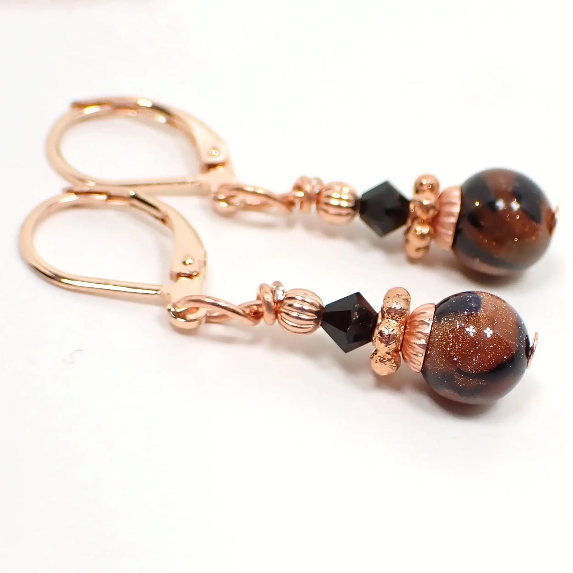 Side view of the handmade goldstone drop earrings. They have bright copper color and rose gold color beads and findings for a copper with hint of pink design. There are faceted black crystal glass bicone beads at the top and small round ball goldstone beads at the bottom. The goldstone beads are marbled with orange glass that has tiny flecks of copper and black.
