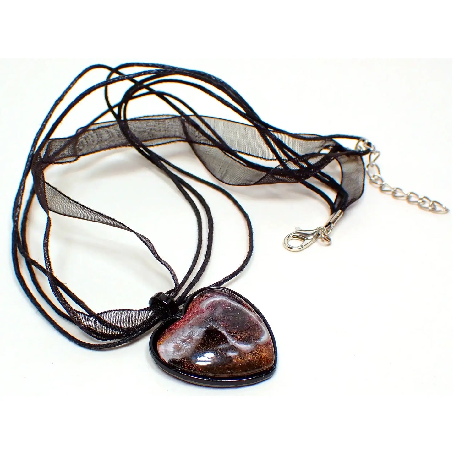 Top view of the handmade resin heart pendant. The necklace part is black and has three strands of faux leather cord with a strand of organza ribbon. There is a lobster clasp and extender chain at the ends. The pendant is a black coated heart shaped setting with a domed resin cab. The resin has shades of iridescent pink with hints of orange depending on the lighting. There is a white abstract frost like formation design through the resin.