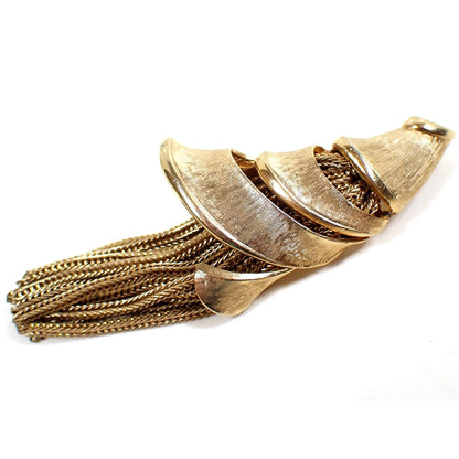 Angled side view of the Mid Century vintage tassel brooch pin. There is a cone shell or horn of plenty type shape wound around the top part of the brooch. There are braided strands of wheat chain coming out of the bottom of the brooch to form the tassel. The metal is gold tone in color and has a matte brushed appearance.