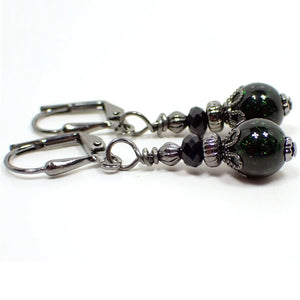 Side view of the small handmade green goldstone earrings. The metal is gunmetal gray in color. There are small faceted black glass crystal beads at the top. The bottom beads are round sphere shaped and are dark green in color with tiny flecks of metallic green for sparkle.