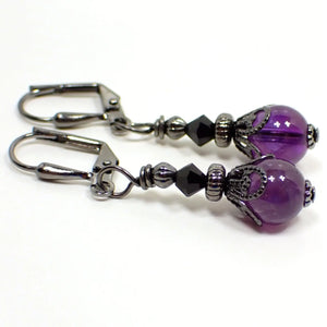 Angled view of the handmade amethyst earrings. The metal is gunmetal plated in color. There are small black glass crystal faceted beads at the top. The gemstone beads on the bottom are round sphere shaped and are dark purple in color.