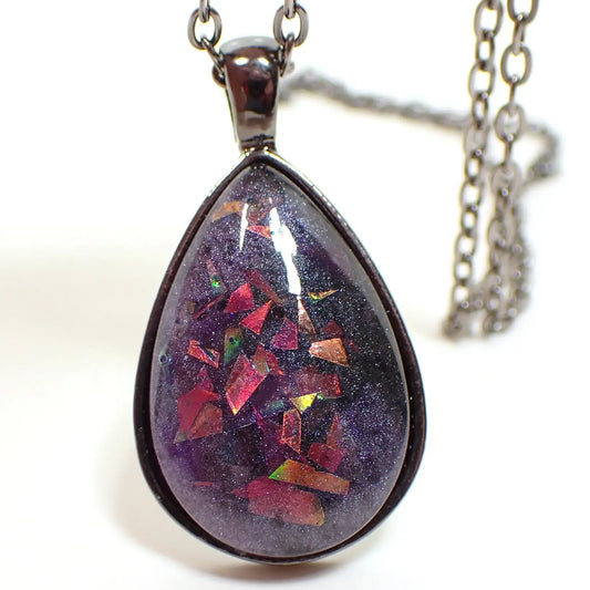 Enlarged front view of the handmade resin pendant necklace with chunky glitter. The metal is gunmetal gray in color. The pendant is teardrop shaped and has a domed resin cab. The resin is pearly purple with chunky pieces of iridescent glitter that flashes with different colors depending on how the light is hitting it.