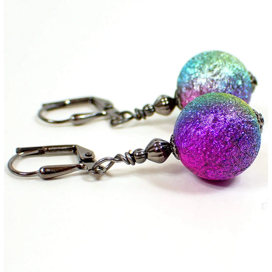 Side view of the handmade rainbow drop earrings. The metal is gunmetal gray in color. There are round ball shaped acrylic beads at the bottom with a textured metallic appearance. The beads have blended shades of purple, blue, green, pink, and yellow.