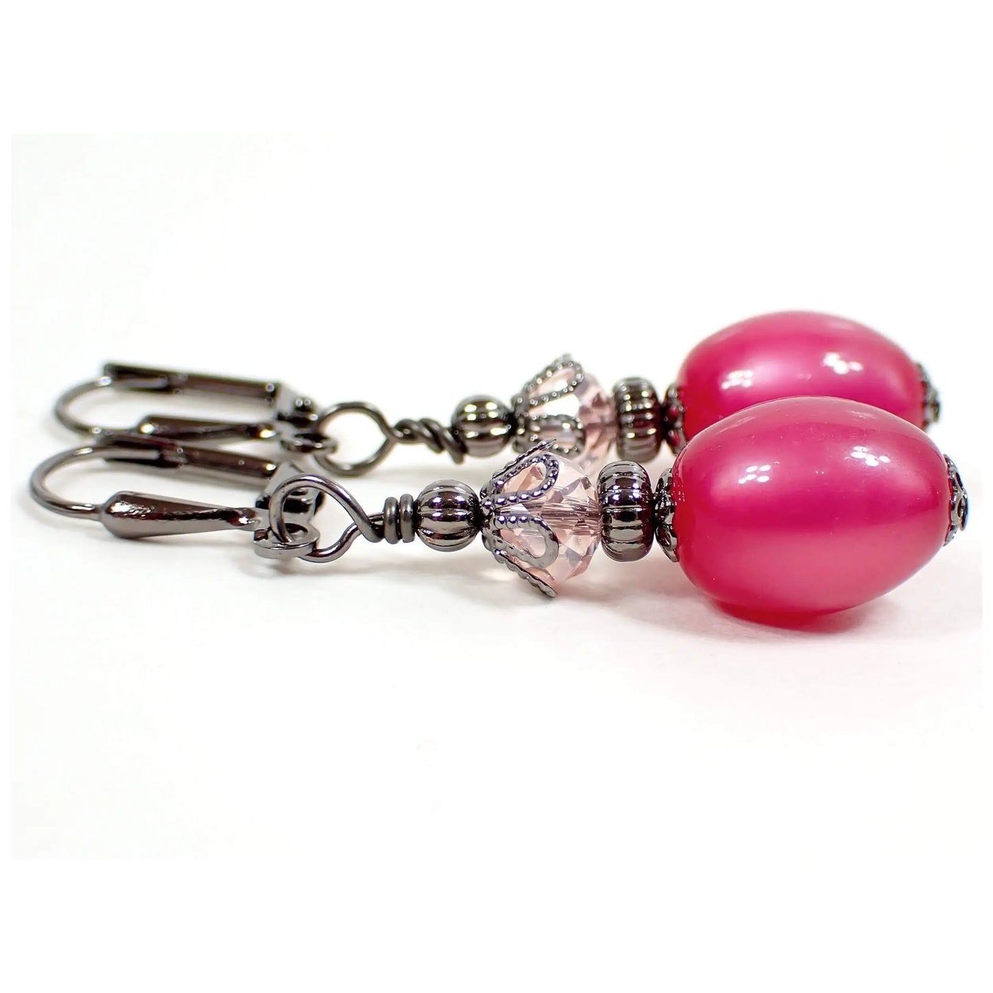 Side view of the handmade drop earrings with vintage moonglow lucite beads. The metal is gunmetal gray in color. There are faceted glass crystal beads at the top in a light pink color. The bottom moonglow lucite beads are raspberry pink in color and are oval shaped. They have an inner glow like appearance when you move around in the light.