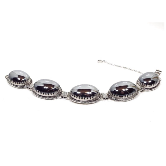 Angled side view of the Mid Century vintage Whiting and Davis link bracelet. It has large oval links with fancy crown like prong settings. The settings each have a large oval metallic gray glass cab to look like hematite. There is a snap lock clasp on the end as well as a safety chain.
