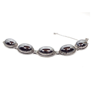 Angled side view of the Mid Century vintage Whiting and Davis link bracelet. It has large oval links with fancy crown like prong settings. The settings each have a large oval metallic gray glass cab to look like hematite. There is a snap lock clasp on the end as well as a safety chain.