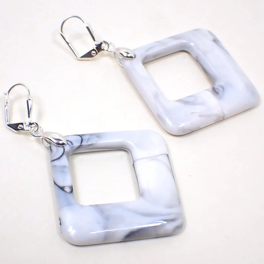 Angled view of the handmade acrylic drop earrings. The metal is silver tone plated in color. They are shaped like squares that hang diagonally and have an opening of the same shape towards the top. They are white with gray and black marbled in.