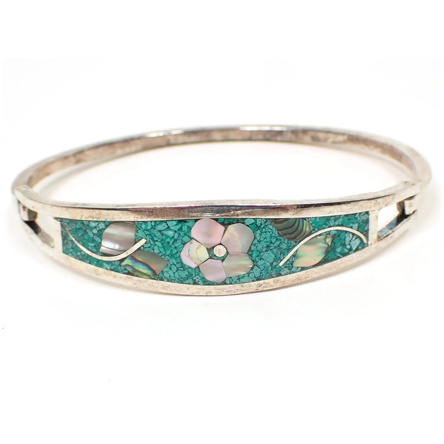 Front view of the retro vintage Mexican floral hinged bangle bracelet. The silver tone metal has a slightly darkened patina from age. The front has tiny pieces of dyed green inlaid stone chips. The flower design has mother of pearl shell pieces for the petals and abalone shell for the leaves.