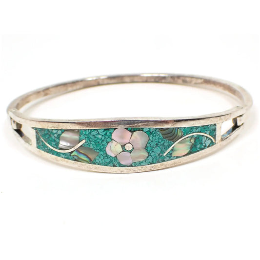 Front view of the retro vintage Mexican floral hinged bangle bracelet. The silver tone metal has a slightly darkened patina from age. The front has tiny pieces of dyed green inlaid stone chips. The flower design has mother of pearl shell pieces for the petals and abalone shell for the leaves.