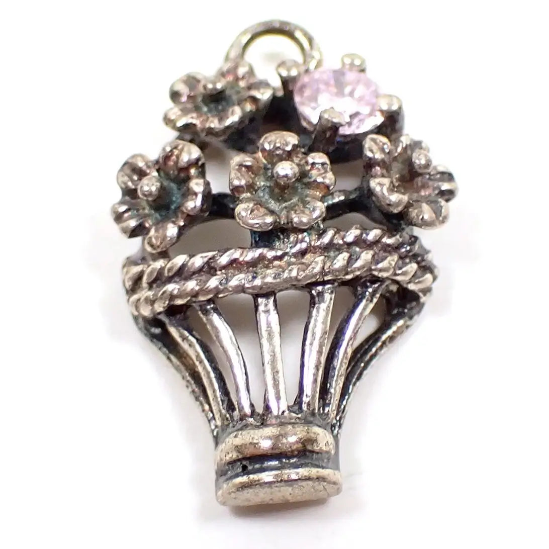 Enlarged view of the retro vintage basket of flowers charm. The bottom part of the charm is shaped like an open basket with a twisted edge at the top. There are four flowers coming out of the top of the basket and a small light pink rhinestone on the top right. The sterling is darkened in color from age and there are some hints of verdigris on the very inside areas of the flowers.