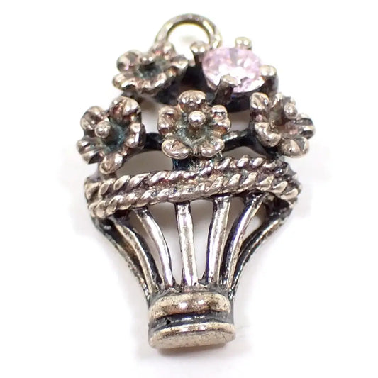 Enlarged view of the retro vintage basket of flowers charm. The bottom part of the charm is shaped like an open basket with a twisted edge at the top. There are four flowers coming out of the top of the basket and a small light pink rhinestone on the top right. The sterling is darkened in color from age and there are some hints of verdigris on the very inside areas of the flowers.