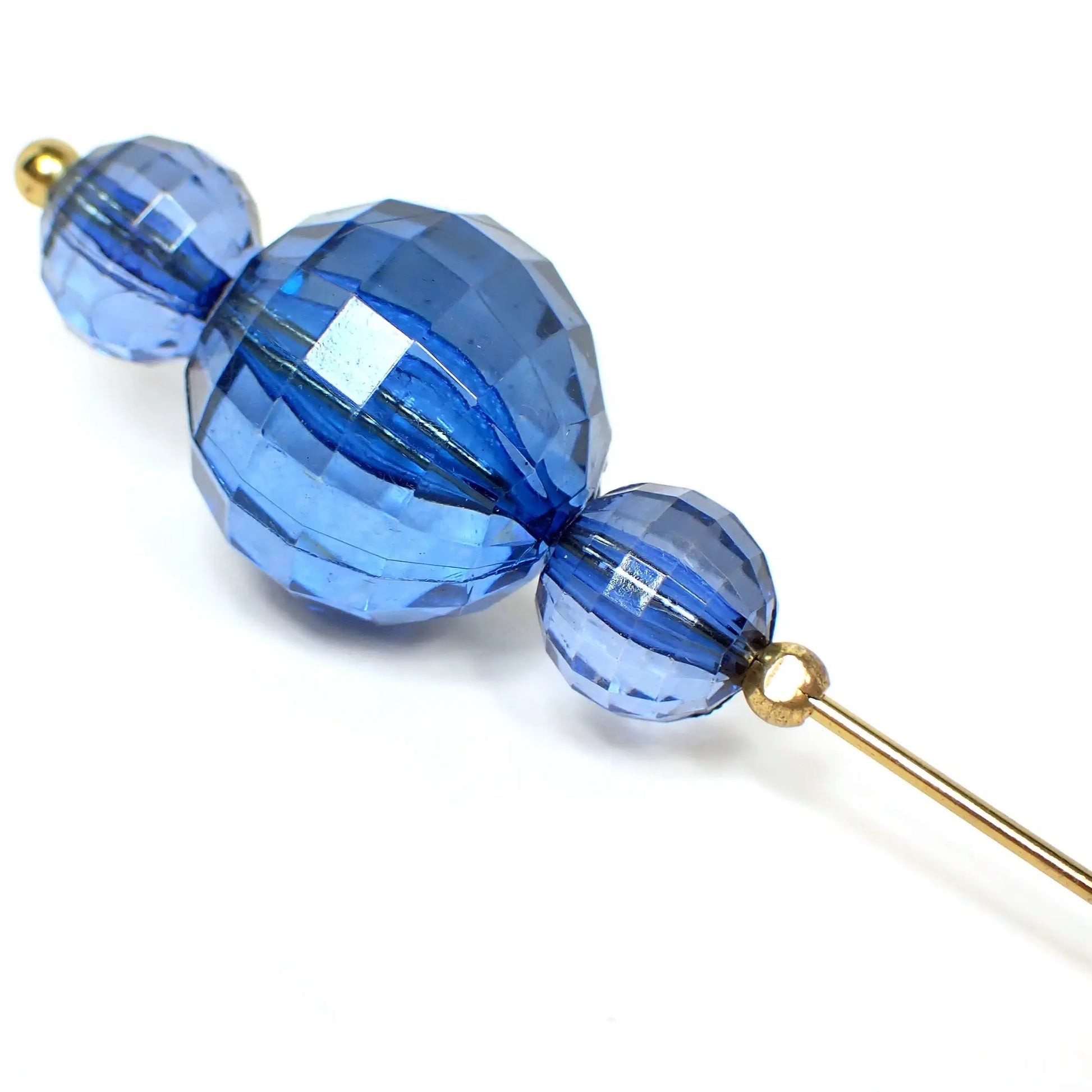 Enlarged view of the top of the Mid Century vintage beaded hat pin. The metal is antiqued gold in color. There are three faceted translucent blue plastic beads. They are all round in shape with the largest bead in the middle. 