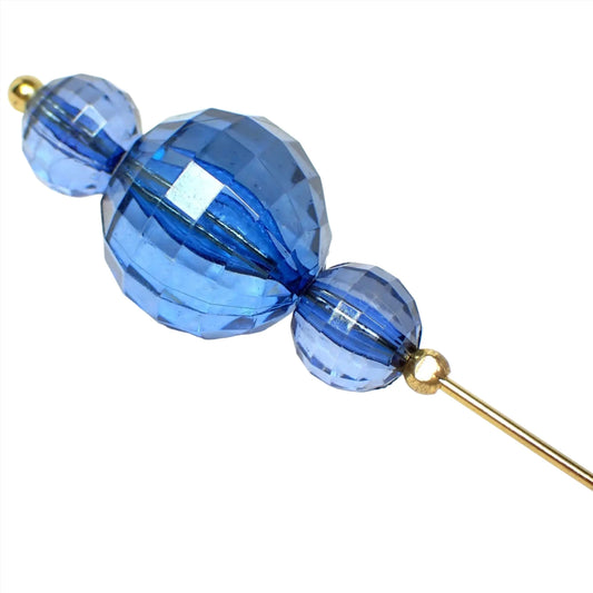 Enlarged view of the top of the Mid Century vintage beaded hat pin. The metal is antiqued gold in color. There are three faceted translucent blue plastic beads. They are all round in shape with the largest bead in the middle. 