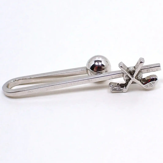 Front view of the retro vintage golfing tie bar. The metal is silver tone in color. There is a squared bar that is curved around to the back to make the tie bar slide on style. The back has a ball end to hold it in place. The front has a design with a golf ball and crossed golf clubs.