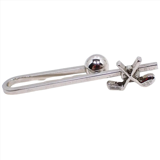 Front view of the retro vintage golfing tie bar. The metal is silver tone in color. There is a squared bar that is curved around to the back to make the tie bar slide on style. The back has a ball end to hold it in place. The front has a design with a golf ball and crossed golf clubs.