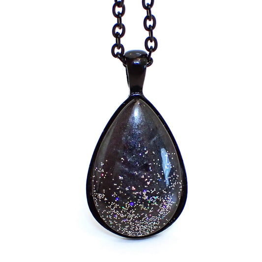 Enlarged front view of the handmade resin pendant necklace. The metal is black coated. The pendant is domed teardrop shapes with pearly dark gray resin. There is holographic glitter scattered around the bottom area of the teardrop for tiny bits of sparkle and flashes of color.