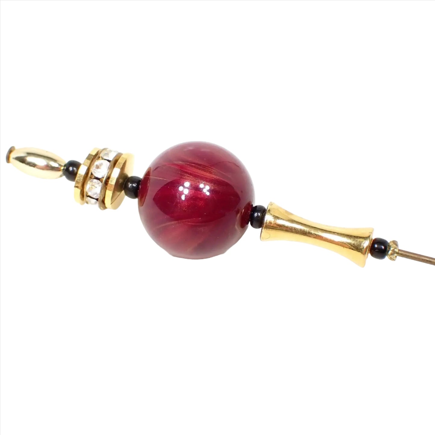 Enlarged angled view of the top of the retro vintage stick pin. The metal is gold tone in color, but the pin has darkened some from age for a more antiqued gold tone appearance. There is a disc shaped bead at the top with clear round rhinestones all the way around it. Underneath that is a round ball shaped lucite bead that has marbled shades of purple and pink for a fuchsia look.