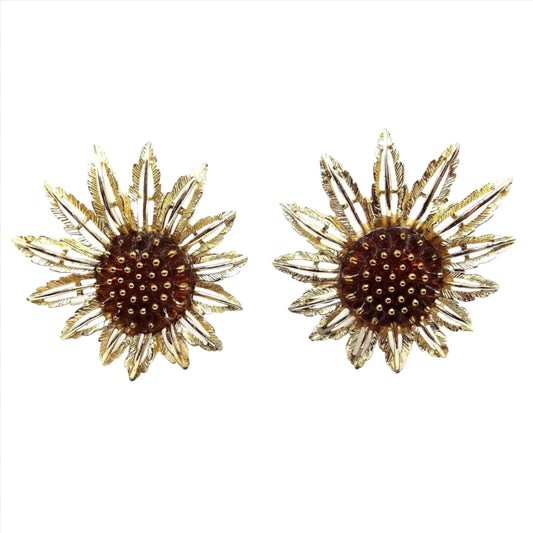 Front view of the Mid Century vintage Sarah Coventry "Starburst" flower earrings. The metal is gold tone in color. There are open cut out style petals with larger ones on one side and shorter on the other. The middle has a dark orange lucite cab with metallic gold painted tips.