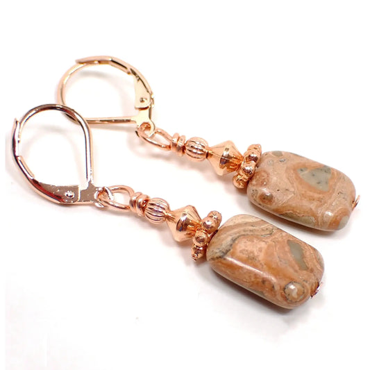 Angled view of the handmade safari jasper earrings. There are lever back earwires at the top in this photo. They are a rose gold in color and the rest of the metal findings are a bright copper plated color. The gemstones are puffy oval shaped and have marbled areas of soft green, brown, peach, and orange.