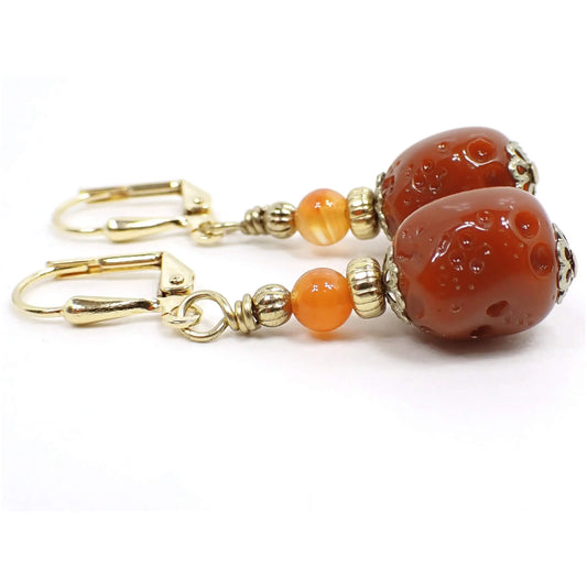 Side view of the handmade drop earrings made with vintage German acrylic beads. The metal is gold plated in color. There vintage acrylic beads at the bottom are burnt orange in color and have a texture pitted nugget like appearance. Small round carnelian gemstone beads were added to the tops of the earrings and are a brighter orange in color with some clear marbled in.