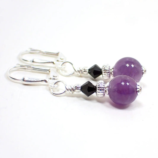 Angled view of the handmade amethyst earrings. The metal is silver tone in color. There are small black glass crystal faceted beads at the top. The gemstone beads on the bottom are round sphere shaped and are purple in color.