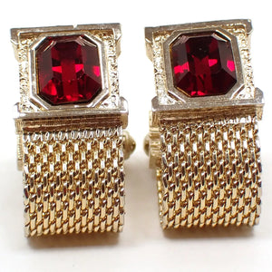 Enlarged front view of the Mid Century vintage Swank wrap around cufflinks. The metal is gold tone plated in color. The top part has a square shape with textured metal around octagon shaped rhinestones in a deep red color. At the bottom is woven metal mesh straps that go around to the back of the cufflinks.