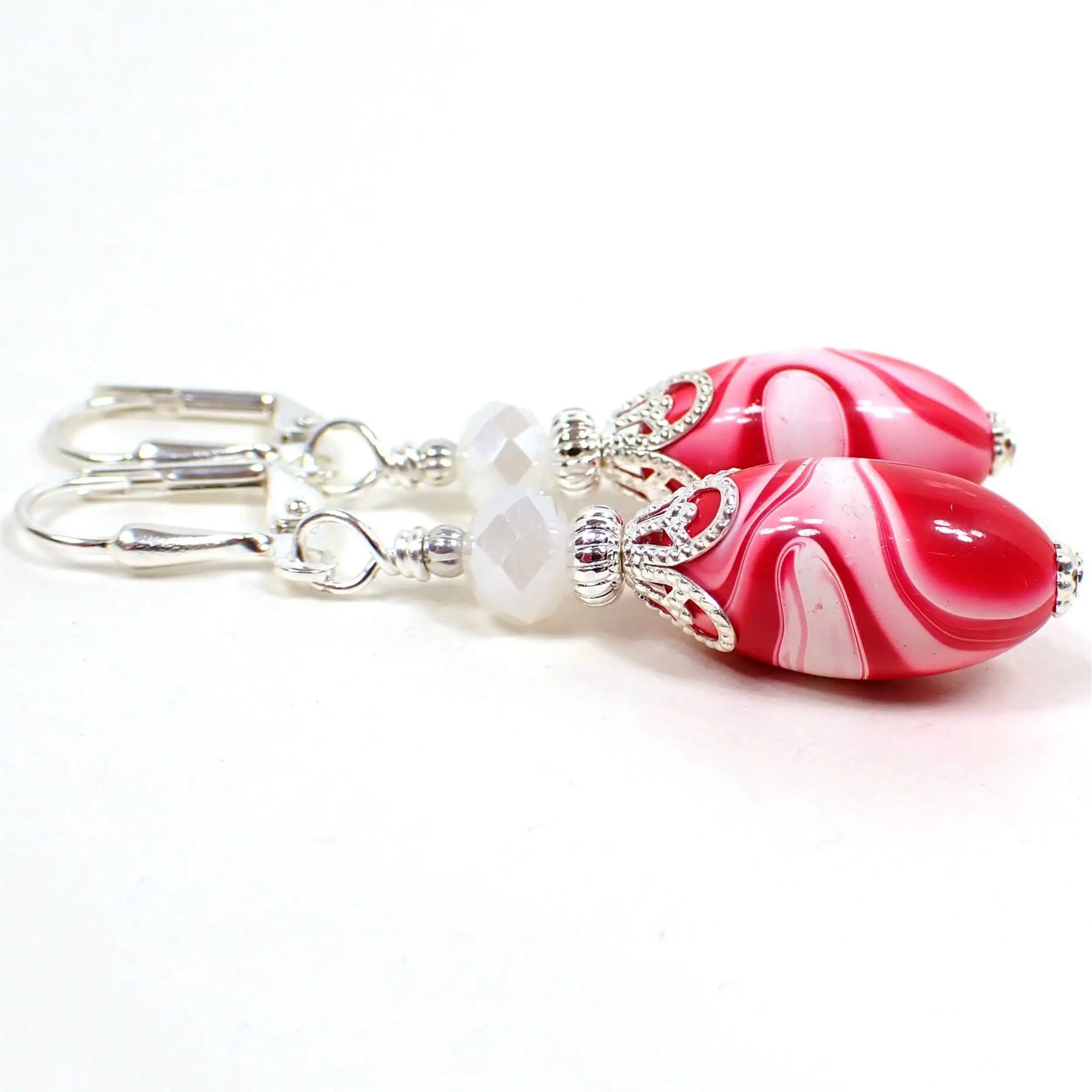 Angled view of the handmade earrings with vintage lucite beads. The metal is silver plated in color. There are faceted opaque pearly white glass beads at the top. The bottom lucite beads are oval shaped and have marbled swirls of red, pink, and white. Each bottom bead has a different pattern from the other for a unique appearance.
